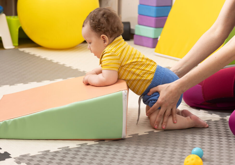 Infant physical therapy session with specialist in rehab center. Little boy patient with muscle weakness doing treatment exercises standing on knees near sport mat